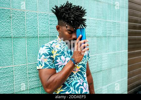 Mittelerwachsener Mann mit locs, die das Mobiltelefon im Stehen halten Gegen die Wand Stockfoto