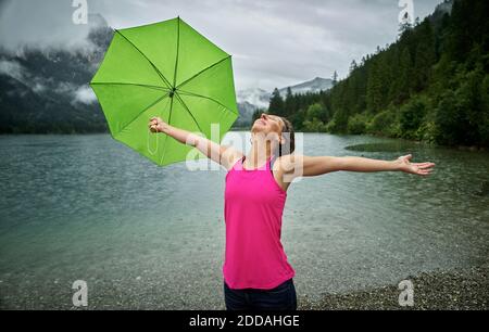 Glückliche Frau, die den Schirm hält, während sie mit ausgestreckten Armen steht Haldensee Seeufer während der Regenzeit Stockfoto