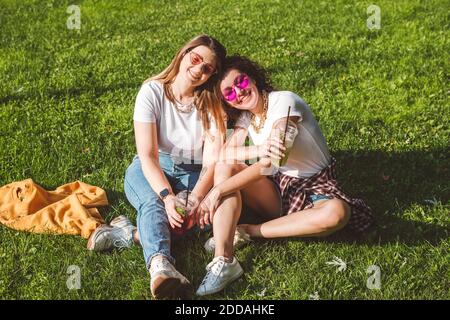 Glückliche Freundinnen sitzen mit Limonade auf Gras im Park An sonnigen Tagen Stockfoto