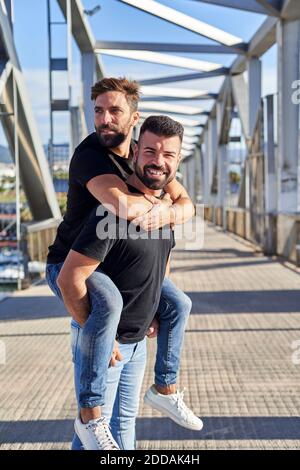 Lächelnd Gay Mann huckbacking Freund auf Fußgängerbrücke während sonnigen Tag Stockfoto