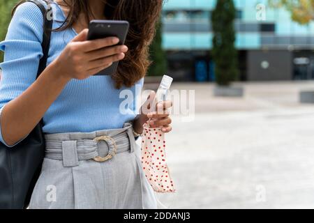 Frau mit Schutzmaske und Desinfektionsmittel mit Smartphone In der Stadt während der COVID-19 Pandemie Stockfoto