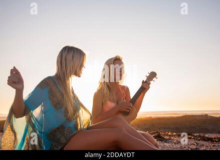 Junge Frau spielt Ukulele für weibliche Freundin am Strand während Sonnenuntergang Stockfoto