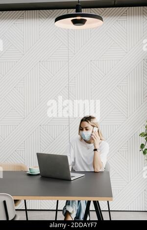 Frau trägt schützende Gesichtsmaske sprechen auf Smartphone, während Laptop im Café benutzen Stockfoto