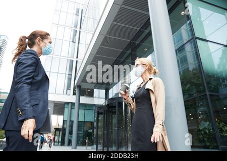 Geschäftsleute, die Gesichtsmaske tragen, reden, während sie mit sozialen Medien stehen Abstand zum Bürogebäude Stockfoto