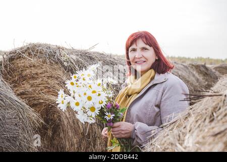 Ältere Frau hält einen Strauß Kamillenblüten, während sie inmitten steht Heuballen Stockfoto