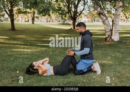 Mann, der Frau dabei hilft, Sit-ups zu machen, während sie auf Gras sitzt Im Hinterhof Stockfoto