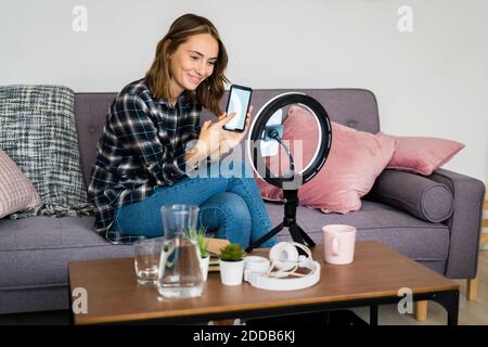 Junge Frau zeigt Handy an Ring-Blitz angeschlossen, während Live-Streaming zu Hause Stockfoto