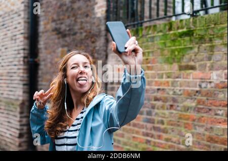 Sorglose Frau mit in-Ear-Kopfhörern, die beim Nehmen die Zunge herausstreckt Selfie über Handy in der Stadt Stockfoto