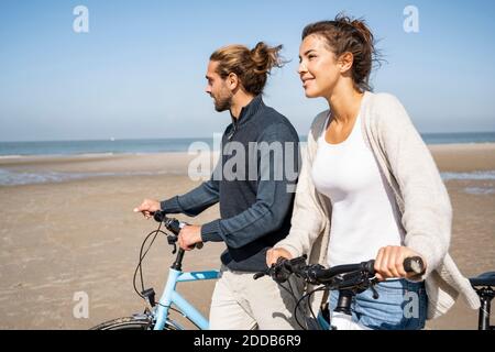 Lächelnde junge Frau, die am Strand mit ihrem Freund und Fahrrädern läuft Gegen klaren Himmel an sonnigen Tag Stockfoto