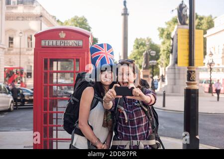 Fröhliche Frau, die Selfie mit einem Freund in der britischen Flagge Hut Gegen rote Telefondose in der Stadt Stockfoto