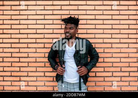 Afrikanischer Mann in Lederjacke, der Musik hört, während er dagegen steht Ziegelwand Stockfoto