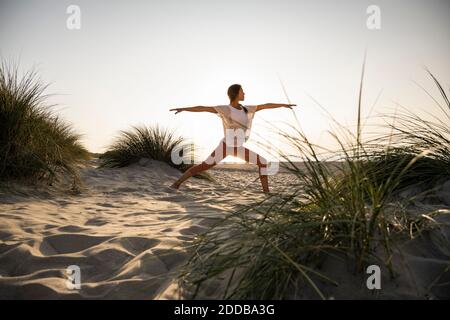 Junge Frau praktiziert Krieger 2 Position Yoga inmitten von Pflanzen an Strand gegen klaren Himmel während des Sonnenuntergangs Stockfoto