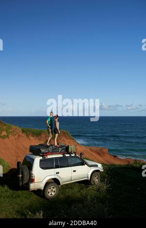 Mittelerwachsenes Paar, das auf dem 4x4-Dach steht, während es sich anschaut Meer gegen blauen Himmel Stockfoto