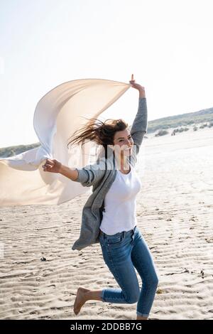 Fröhliche Frau läuft, während sie die Decke am Strand bei Sonnenschein hält Tag Stockfoto
