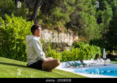 Männlicher Tourist meditiert während er Yoga auf Gras in der Nähe von Schwimmen praktiziert Pool im Gesundheitsbereich an sonnigen Tagen Stockfoto