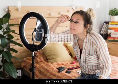 Weibliche Influencer Anwendung Mascara während vlogging durch Smartphone sitzen Im Schlafzimmer zu Hause Stockfoto