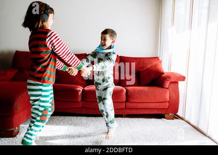 Bruder und Schwester mit Kopfhörern, die die Hände halten, während sie dagegen tanzen Sofa im Wohnzimmer Stockfoto