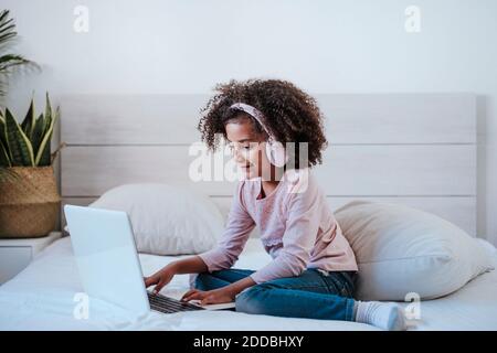 Nettes Mädchen mit Laptop und Kopfhörer tragen, während im Sitzen Schlafzimmer zu Hause Stockfoto