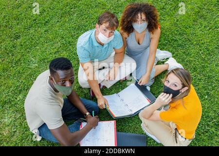 Schüler tragen Schutzmaske studieren, während sie zusammen auf Gras sitzen Auf dem Universitätsgelände Stockfoto