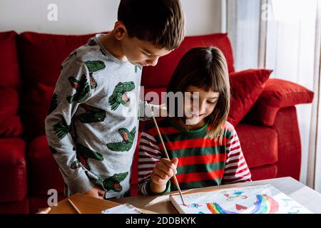 Lächelnde Schwester Malerei Regenbogen von Bruder im Wohnzimmer an Zu Hause Stockfoto