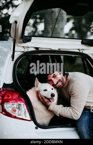 Lächelnder Mann umarmt Hund, während er im Kofferraum sitzt Wald Stockfoto