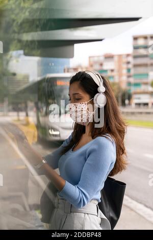 Frau in Gesichtsmaske trägt Kopfhörer während der Verwendung von Smartphone Am Busbahnhof während der COVID-19 Stockfoto