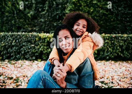 Lächelndes Mädchen umarmt Mutter von hinten, während sie im Park sitzt Stockfoto