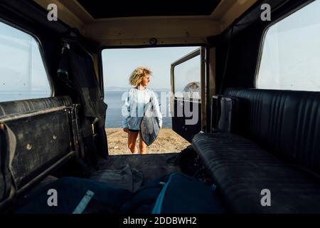 Frau hält Surfbrett, während sie mit dem Auto gegen das Meer steht Strand Stockfoto