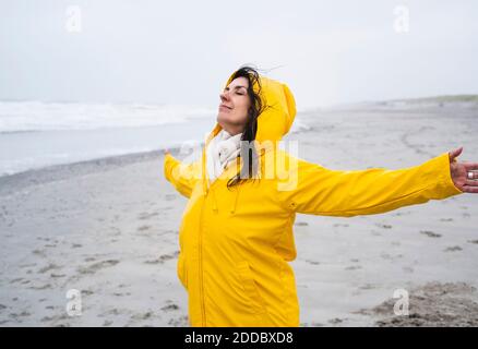 Reife Frau im gelben Regenmantel stehend mit ausgestreckten Armen Strand Stockfoto