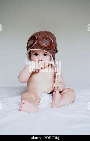 Baby Junge mit Finger im Mund trägt Fliegermütze sitzend Auf dem Bett zu Hause Stockfoto