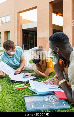 Studentin mit Freunden, die eine Sicherheitsmaske tragen, während sie über das Studium lernt Gras auf dem Universitätscampus Stockfoto