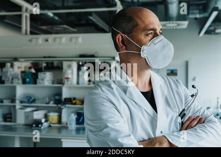 Männlicher Wissenschaftler trägt Gesichtsmaske wegschauen, während mit stehen Arme im Labor gekreuzt Stockfoto
