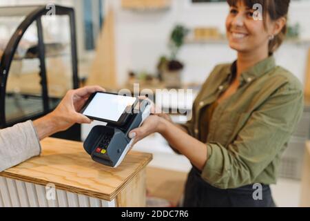 Kurze Hand des Kunden, der kontaktloses Bezahlen mit Smartphone macht An die Besitzerin beim Auschecken im Café Stockfoto