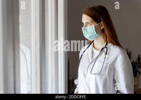 Junge Ärztin in Schutzmaske Blick durch Fenster Während COVID-19 Stockfoto