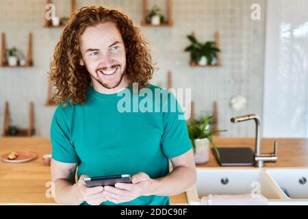 Lächelnder Mann, der wegschaut, während er in der Küche ein digitales Tablet benutzt Von Studio-Apartment Stockfoto