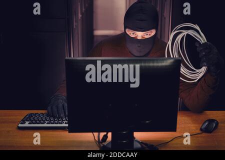 Ein Mann in einer schwarzen Maske mit Drähten in der Hand sitzt an einem Computer in einem Serverraum. Konzept von Datenintegritätsproblemen und Hackerangriffen Stockfoto