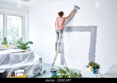 Junge Frau malt Wand mit Schmerzrolle, während sie auf steht Leiter zu Hause Stockfoto