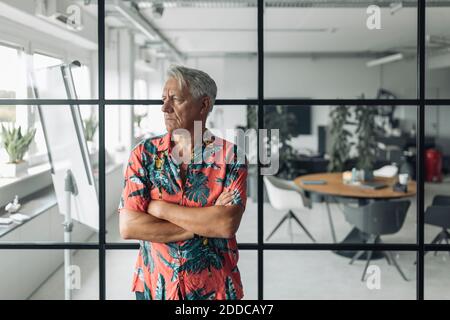 Geschäftsmann mit gekreuzten Armen, der im Büro gegen eine Glaswand steht Stockfoto