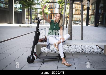 Lächelnde Frau mittleren Erwachsenen Musik hören und Selfie während Sitzen auf der Bank in der Stadt Stockfoto