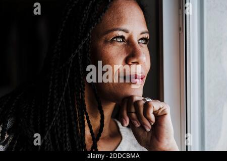 Reife Frau mit der Hand auf dem Kinn, die durch das Fenster im Stehen schaut Zu Hause Stockfoto