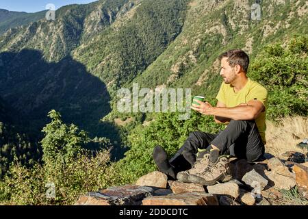 Mann, der Kaffee trinkt und wegschaut, während er auf Stein sitzt Wald an sonnigen Tagen Stockfoto