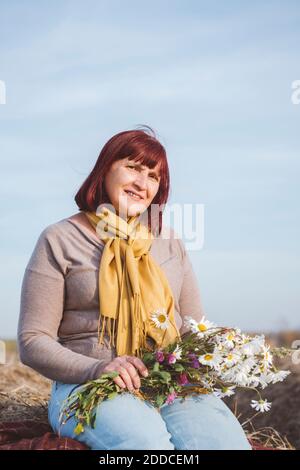 Ältere Frau sitzt auf Heuballen, während sie Kamillenblüten hält Stockfoto