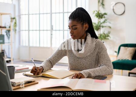 Studentin, die im Buch auf dem Tisch schreibt, während sie sitzt Zu Hause Stockfoto