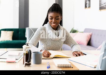 Lächelnde Frau schreibt im Buch, während sie im Wohnzimmer sitzt Zu Hause Stockfoto