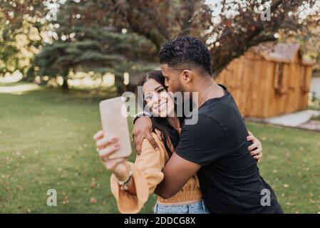 Mann küsst Frau, die Selfie durch Handy während des Stehens Im Hinterhof Stockfoto