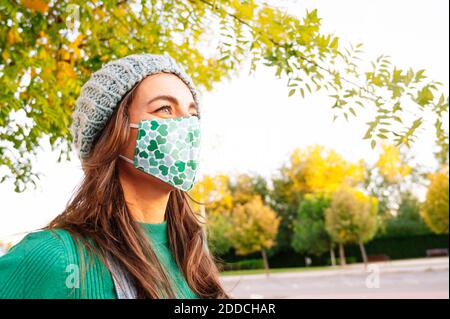 Reife Frau schaut weg, während das Tragen von schützenden Gesichtsmaske stehen Im öffentlichen Park an sonnigen Tag Stockfoto