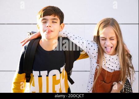 Bruder und Schwester ragen aus der Zunge, während sie mit dem Arm standen Um gegen die Wand an sonnigen Tag Stockfoto