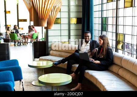 Lächelnde männliche und weibliche Geschäftsleute diskutieren über Laptop, während Sitzen auf dem Sofa in der modernen Bar Stockfoto