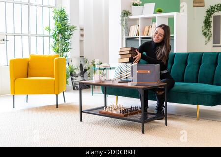Lächelnde junge Frau, die Bücher in der Schachtel auf dem Couchtisch sammelt Im Loft-Apartment Stockfoto