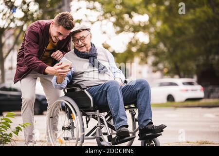 Älterer Mann im Rollstuhl und sein erwachsener Enkel schauen zusammen Auf dem Smartphone